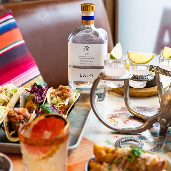 Lalo Tequila bottle, shot glasses with lime, tacos, and a grapefruit cocktail on a table.