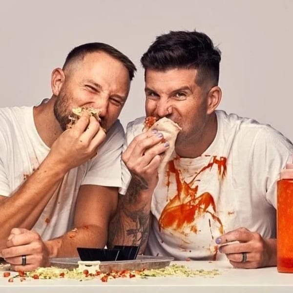 Two men in white shirts, stained with red sauce, happily eating messy tacos.