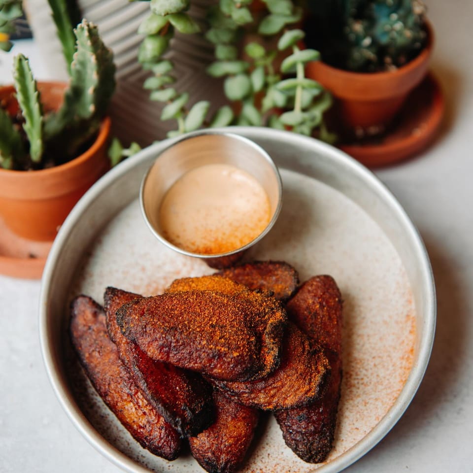 Crispy plantain slices dusted with chili powder and a side of creamy sauce, next to potted plants.