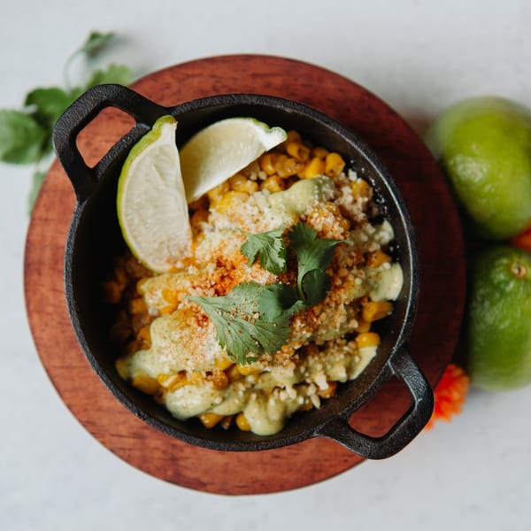 Elote in a black cast iron dish with lime wedges, cilantro, and cotija cheese.