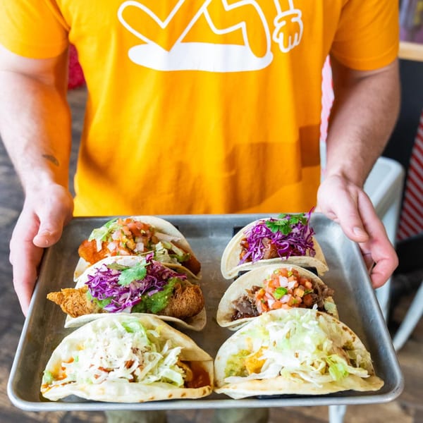 A person in a yellow shirt holding a tray with six different, colorful Hombre y Lobo tacos.