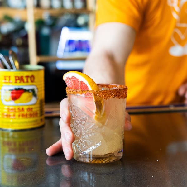 A hand holding a cocktail with a chili-lime rim and grapefruit slice, next to a yellow El Pato can.