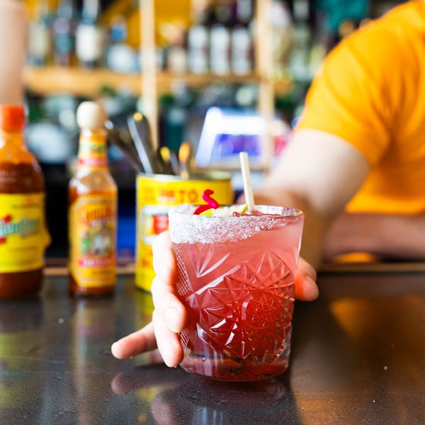 A bartender's hand serves a vibrant red, salt-rimmed cocktail with a straw and garnish.
