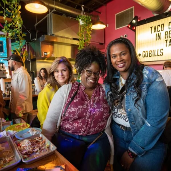 Three smiling women pose in a vibrant Mexican restaurant with food trays and a "Taco 'bout it" sign.