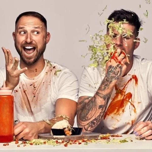 Two men in white shirts covered in hot sauce and lettuce, one laughing, the other eating a taco.