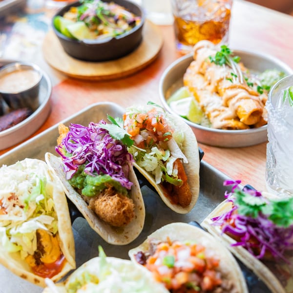 A metal tray holds several vibrant tacos with various toppings, alongside other Mexican dishes and drinks.