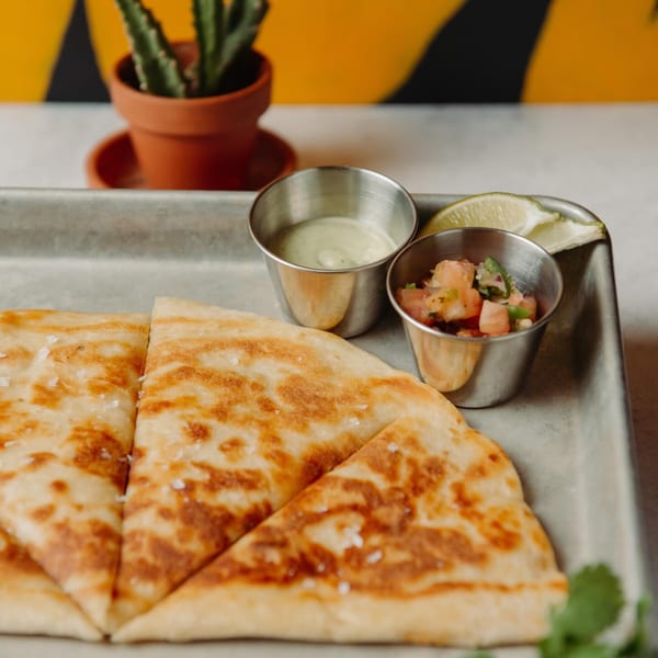 Golden-brown quesadilla slices with salsa, crema, and lime on a metal tray, with a cactus in the background.