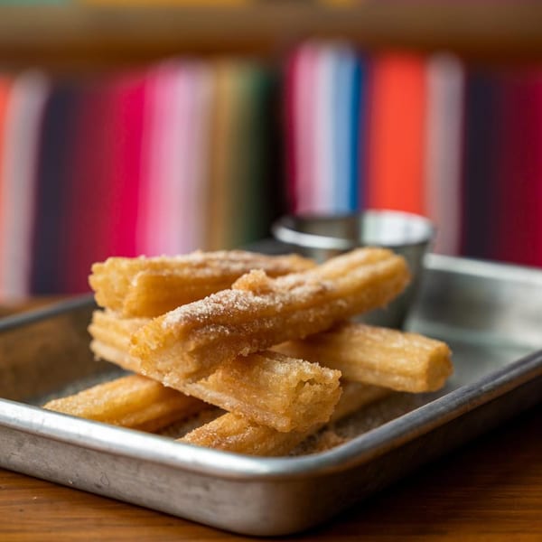 Golden churros dusted with sugar on a metal tray, with a small cup of dipping sauce and colorful background.